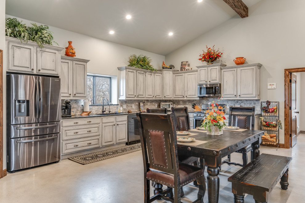 Mid-sized country l-shaped eat-in kitchen in Austin with an undermount sink, raised-panel cabinets, grey cabinets, granite benchtops, grey splashback, stone tile splashback, stainless steel appliances, concrete floors, grey floor, black benchtop and vaulted.