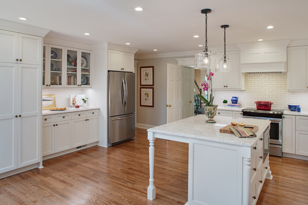 Photo of a large traditional l-shaped eat-in kitchen in Atlanta with a single-bowl sink, flat-panel cabinets, white cabinets, quartz benchtops, white splashback, subway tile splashback, stainless steel appliances, medium hardwood floors, with island, brown floor and white benchtop.