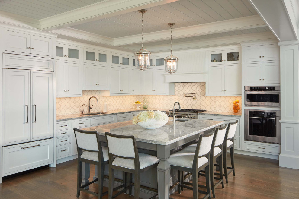 Large beach style l-shaped open plan kitchen in Baltimore with an undermount sink, shaker cabinets, white cabinets, white splashback, stone tile splashback, stainless steel appliances, medium hardwood floors, with island, brown floor, grey benchtop and coffered.