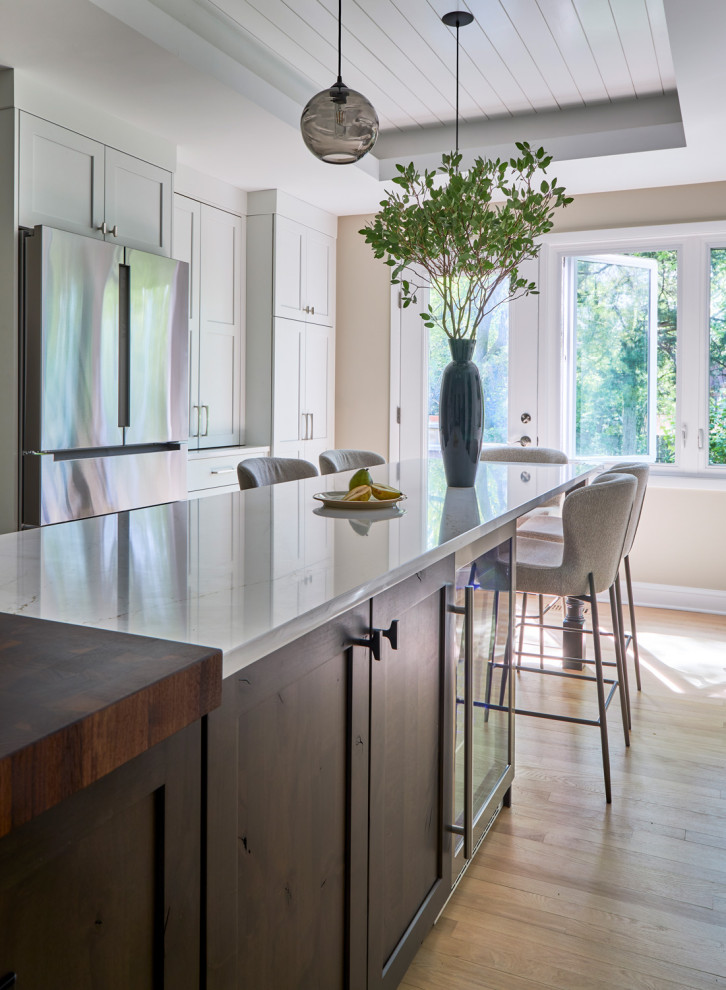 Example of a large transitional u-shaped light wood floor, brown floor and coffered ceiling eat-in kitchen design in Chicago with a farmhouse sink, shaker cabinets, white cabinets, quartz countertops, white backsplash, subway tile backsplash, stainless steel appliances, an island and multicolored countertops