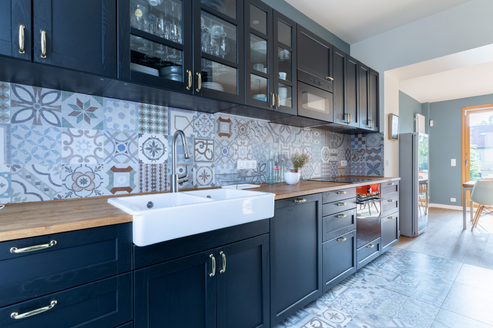 Mid-sized contemporary single-wall open plan kitchen in Paris with a farmhouse sink, black cabinets, wood benchtops, cement tile splashback, black appliances, cement tiles, with island, multi-coloured floor, shaker cabinets, multi-coloured splashback and brown benchtop.