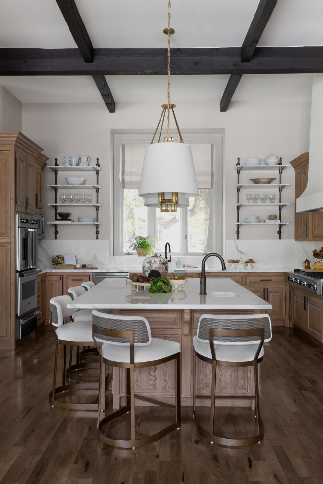 This is an example of a traditional u-shaped kitchen in Austin with an undermount sink, raised-panel cabinets, medium wood cabinets, white splashback, stone slab splashback, stainless steel appliances, dark hardwood floors, with island, brown floor, white benchtop and exposed beam.
