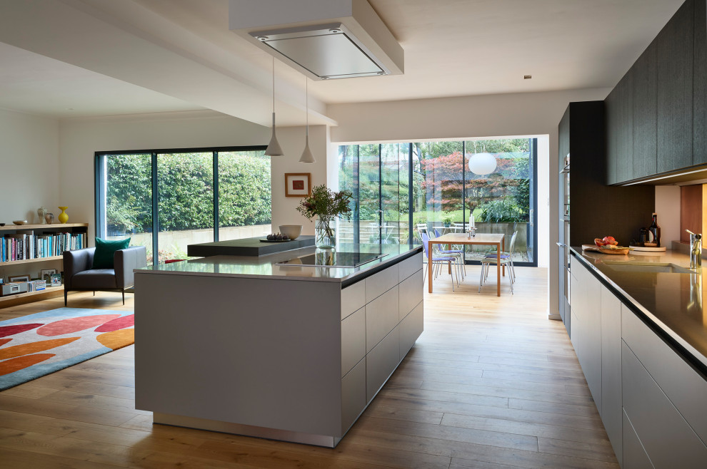 Photo of an expansive modern grey and black single-wall open plan kitchen in London with an integrated sink, flat-panel cabinets, grey cabinets, quartz worktops, multi-coloured splashback, glass sheet splashback, black appliances, light hardwood flooring and an island.