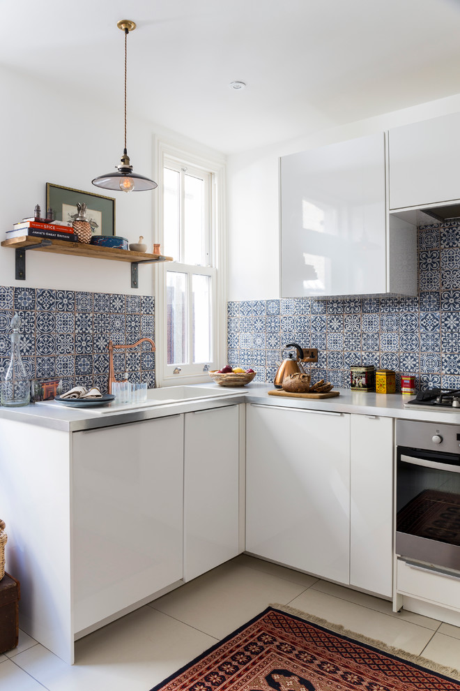 Small eclectic u-shaped separate kitchen in London with a drop-in sink, flat-panel cabinets, white cabinets, black splashback, ceramic splashback, stainless steel appliances, ceramic floors, no island and stainless steel benchtops.