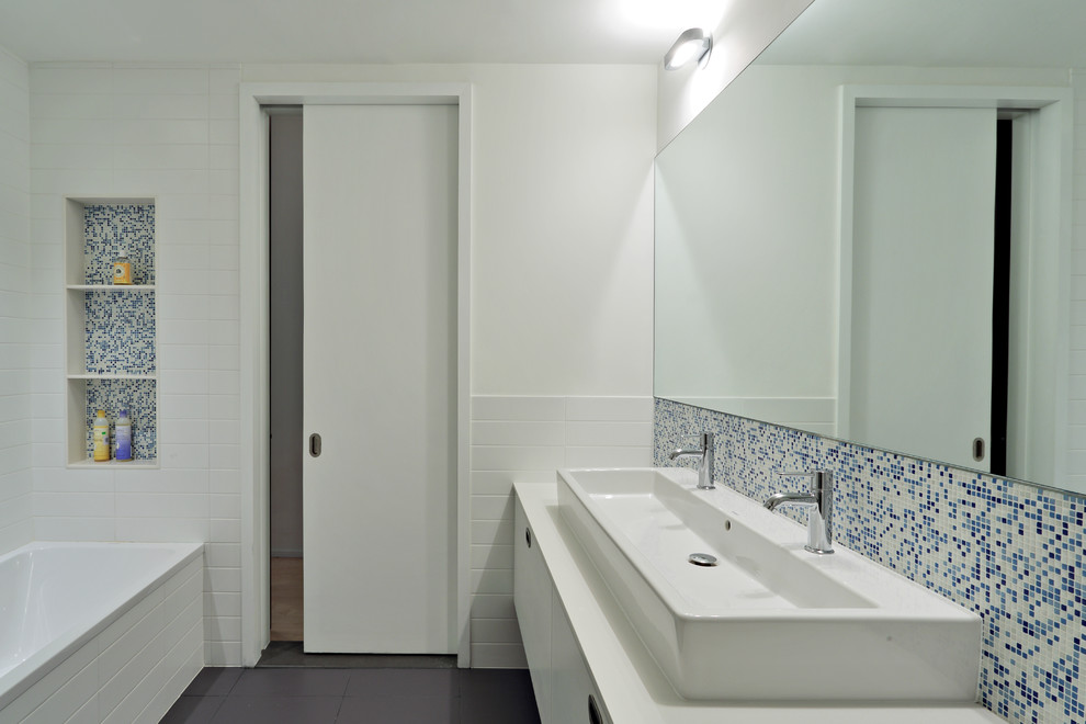 Design ideas for a large modern master bathroom in New York with flat-panel cabinets, an alcove tub, blue tile, white tile, mosaic tile, white walls, a trough sink, white cabinets, an alcove shower, porcelain floors and solid surface benchtops.