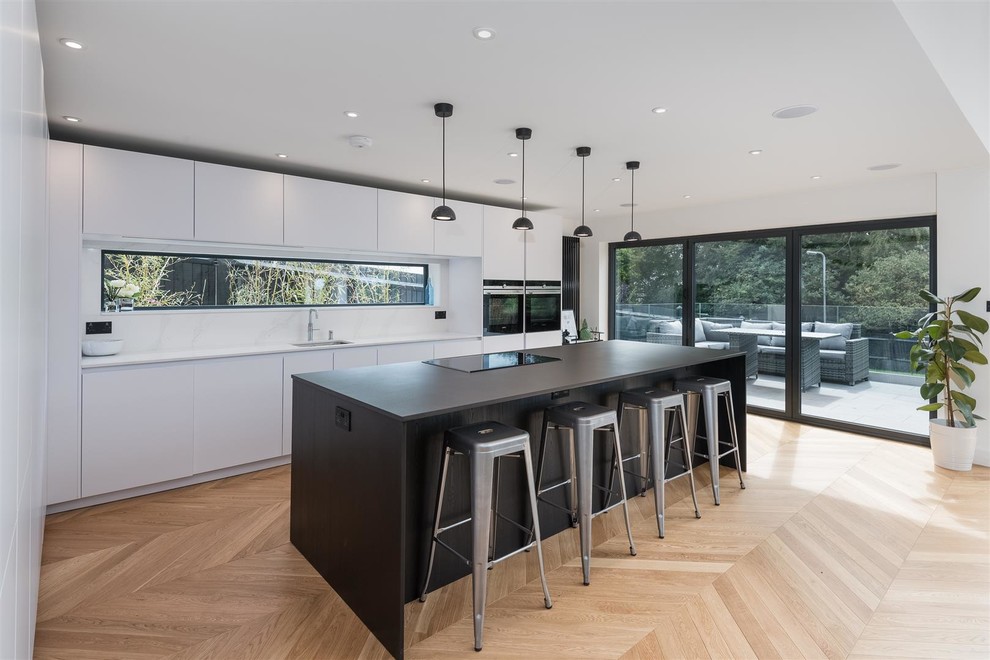 Design ideas for a mid-sized contemporary l-shaped kitchen in Sussex with a single-bowl sink, flat-panel cabinets, window splashback, black appliances, light hardwood floors, with island and white benchtop.