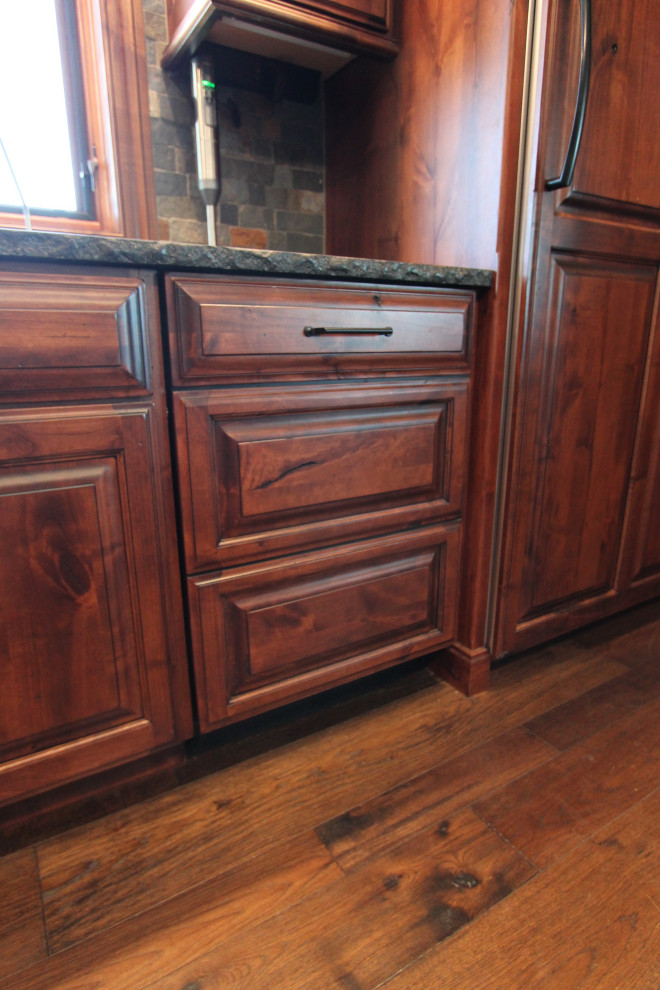 Example of a large mountain style medium tone wood floor and brown floor kitchen design with raised-panel cabinets, medium tone wood cabinets, granite countertops, multicolored backsplash, stone tile backsplash, paneled appliances, an island and black countertops
