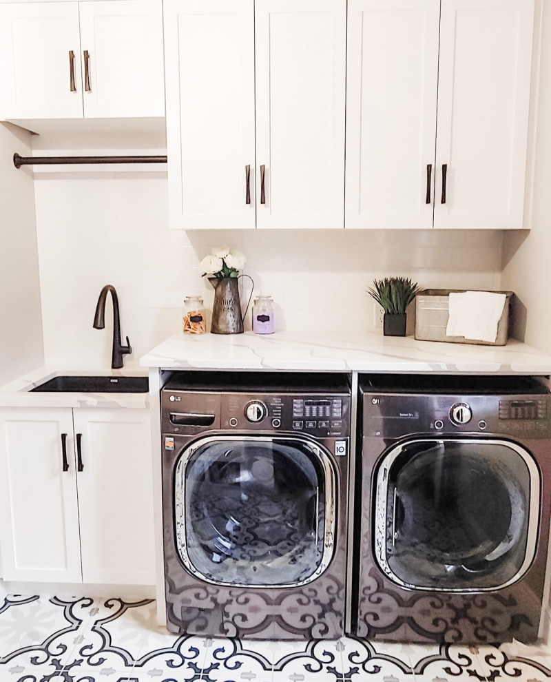 Photo of a mid-sized transitional single-wall utility room with an undermount sink, shaker cabinets, white cabinets, quartz benchtops, white walls, ceramic floors, a side-by-side washer and dryer, multi-coloured floor, white benchtop, white splashback and ceramic splashback.