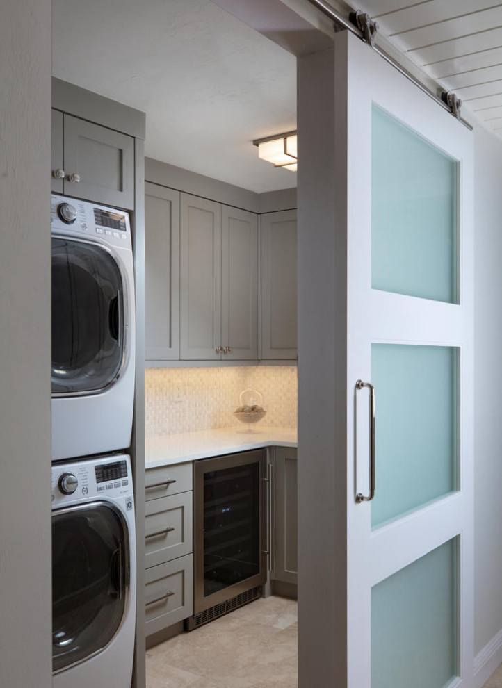 Photo of a small beach style l-shaped dedicated laundry room in Miami with an undermount sink, shaker cabinets, grey cabinets, quartz benchtops, white splashback, ceramic splashback, grey walls, travertine floors, a stacked washer and dryer, beige floor and white benchtop.