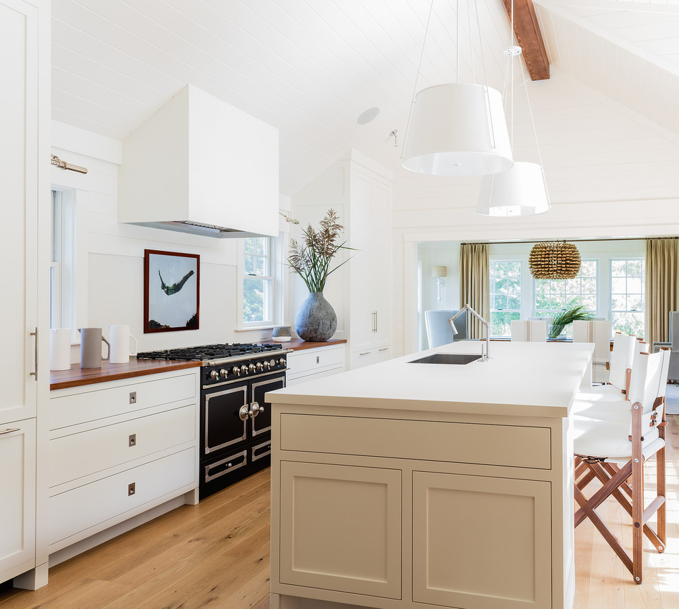 Inspiration for a transitional light wood floor open concept kitchen remodel in Boston with an undermount sink, shaker cabinets, white cabinets, wood countertops, white backsplash, black appliances and an island