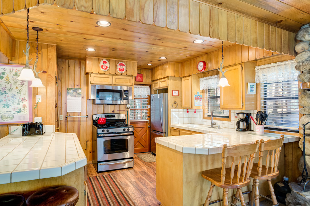 Example of a mountain style medium tone wood floor kitchen design with an undermount sink, recessed-panel cabinets, light wood cabinets, tile countertops, white backsplash, stainless steel appliances and a peninsula