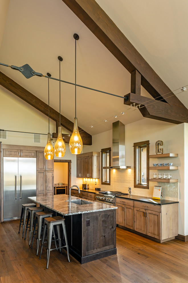 Mountain style medium tone wood floor and brown floor kitchen photo with shaker cabinets, light wood cabinets, an island, an undermount sink, white backsplash, stainless steel appliances and black countertops
