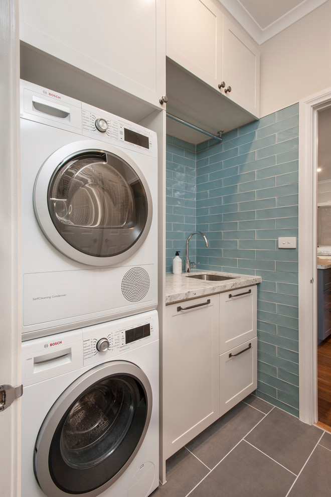 This is an example of a small traditional single-wall dedicated laundry room in Melbourne with an undermount sink, shaker cabinets, beige cabinets, granite benchtops, blue walls, porcelain floors, a stacked washer and dryer, grey floor and white benchtop.