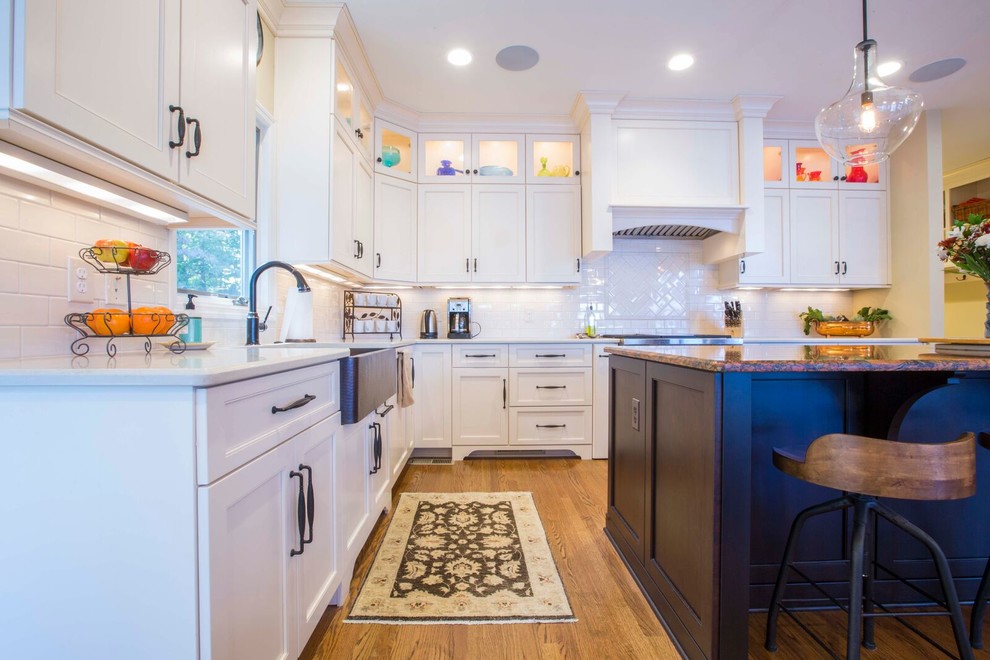 Inspiration for a large transitional l-shaped light wood floor and brown floor enclosed kitchen remodel with a farmhouse sink, shaker cabinets, white cabinets, solid surface countertops, white backsplash, porcelain backsplash, stainless steel appliances and an island