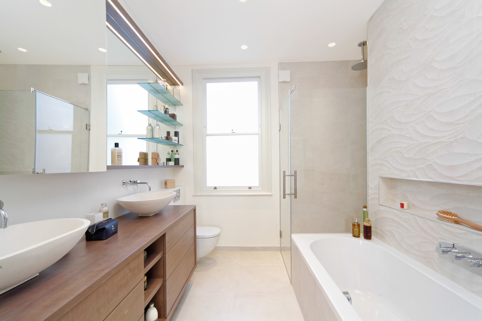Photo of a mid-sized contemporary master bathroom in London with a console sink, flat-panel cabinets, medium wood cabinets, wood benchtops, a drop-in tub, an alcove shower, a wall-mount toilet, beige tile, porcelain tile, beige walls and porcelain floors.