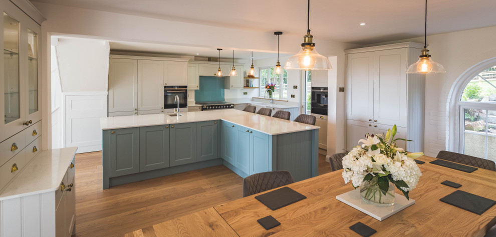 Photo of a large contemporary grey and teal l-shaped open plan kitchen in Devon with a belfast sink, shaker cabinets, turquoise cabinets, quartz worktops, blue splashback, glass sheet splashback, black appliances, light hardwood flooring, an island, brown floors, white worktops and feature lighting.
