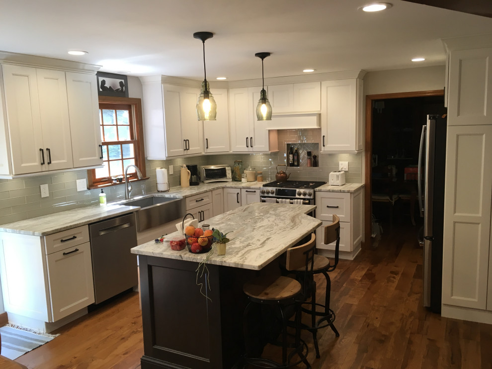 Example of a mid-sized transitional u-shaped medium tone wood floor and brown floor eat-in kitchen design with a farmhouse sink, recessed-panel cabinets, white cabinets, granite countertops, gray backsplash, glass tile backsplash, stainless steel appliances, an island and gray countertops