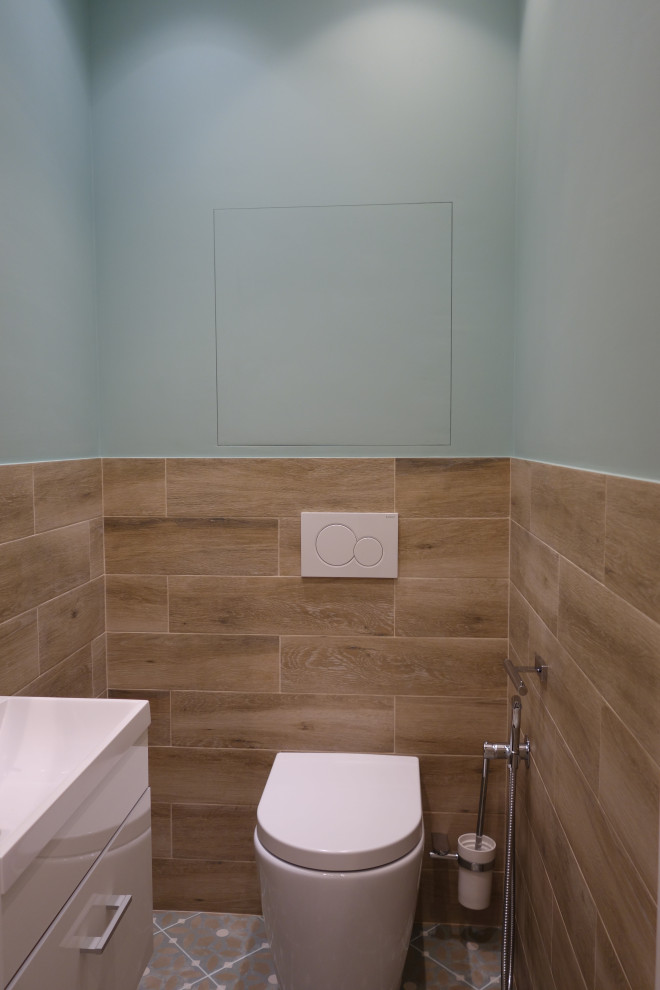 Bild på ett mellanstort maritimt toalett, med släta luckor, vita skåp, en vägghängd toalettstol, brun kakel, porslinskakel, blå väggar, klinkergolv i porslin, ett nedsänkt handfat och beiget golv