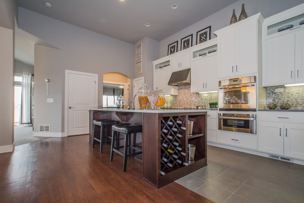 This is an example of a transitional l-shaped eat-in kitchen in Denver with white cabinets, solid surface benchtops, multi-coloured splashback, mosaic tile splashback, stainless steel appliances, with island, an undermount sink, shaker cabinets and medium hardwood floors.