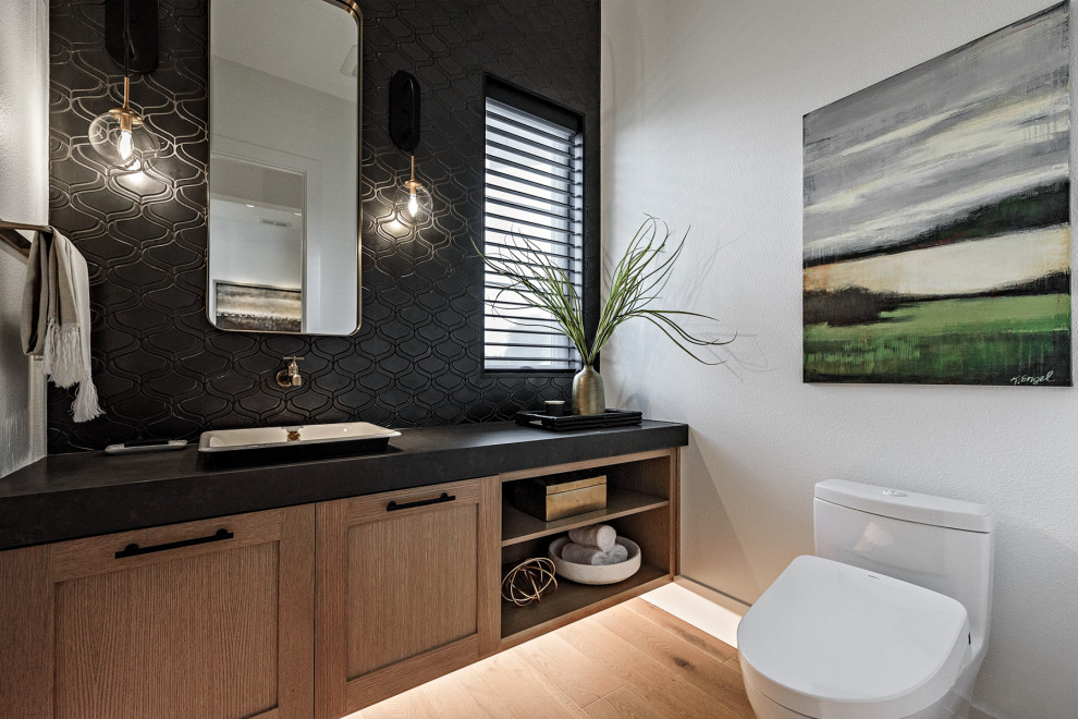 This is an example of a country powder room in Portland with shaker cabinets, light wood cabinets, a one-piece toilet, black tile, white walls, light hardwood floors, a vessel sink, beige floor and black benchtops.
