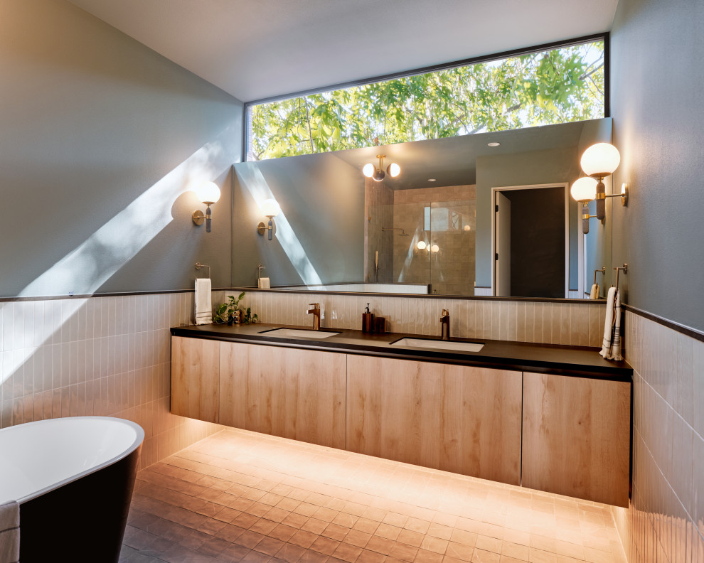 This is an example of a medium sized midcentury ensuite bathroom in Austin with light wood cabinets, a freestanding bath, a shower/bath combination, a two-piece toilet, beige tiles, porcelain tiles, beige walls, porcelain flooring, a submerged sink, solid surface worktops, black floors, a hinged door, black worktops, double sinks, a floating vanity unit, wainscoting and flat-panel cabinets.