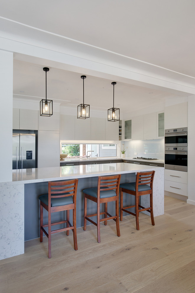 This is an example of a large modern l-shaped open plan kitchen in Sydney with an undermount sink, flat-panel cabinets, white cabinets, quartz benchtops, white splashback, glass sheet splashback, stainless steel appliances, light hardwood floors, with island and grey benchtop.