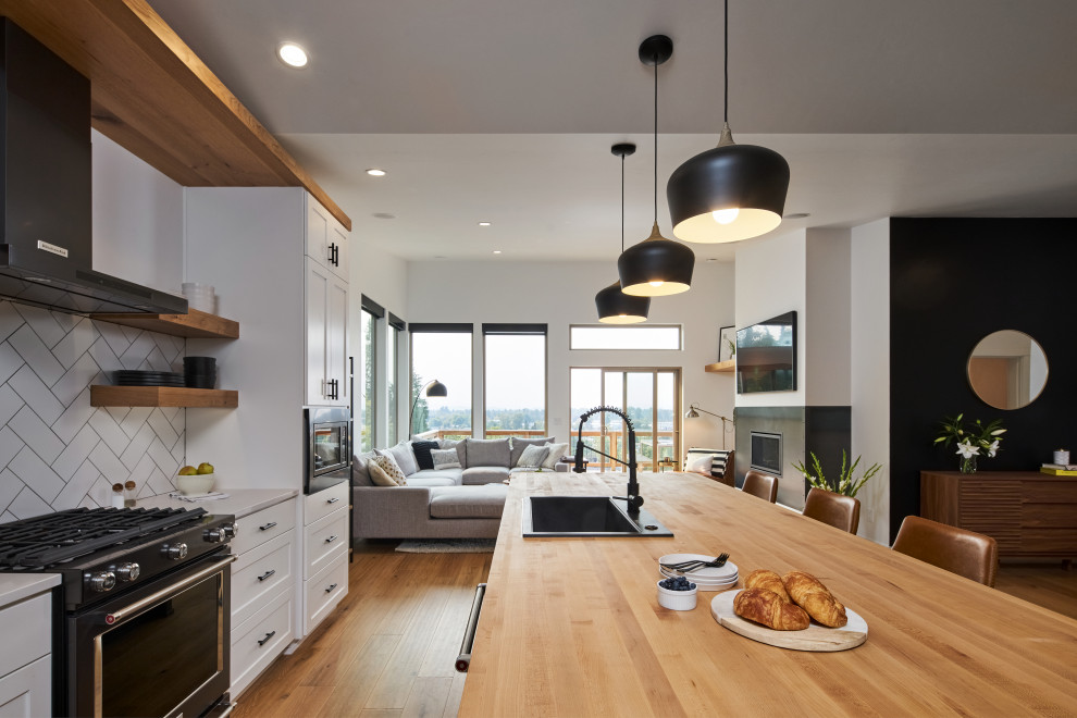 Large modern single-wall open plan kitchen in Other with a drop-in sink, shaker cabinets, white cabinets, wood benchtops, white splashback, cement tile splashback, black appliances, medium hardwood floors, with island, brown floor, brown benchtop and vaulted.