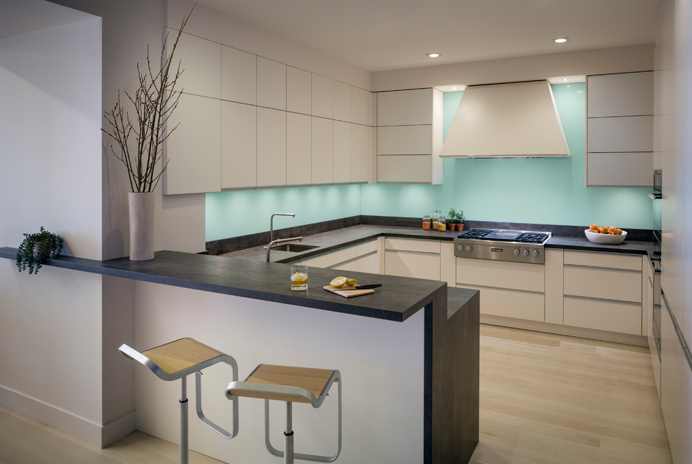 Photo of a mid-sized contemporary u-shaped kitchen in San Francisco with an undermount sink, flat-panel cabinets, blue splashback, stainless steel appliances, light hardwood floors, a peninsula, beige floor, black benchtop, beige cabinets, soapstone benchtops and glass sheet splashback.