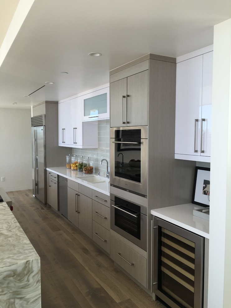 Design ideas for a large contemporary galley open plan kitchen in Los Angeles with an undermount sink, flat-panel cabinets, white cabinets, grey splashback, glass tile splashback, stainless steel appliances, medium hardwood floors, with island and onyx benchtops.