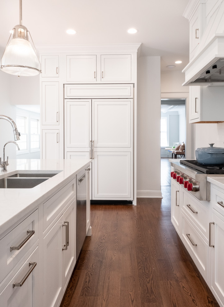 This is an example of a mid-sized transitional l-shaped eat-in kitchen in New York with a double-bowl sink, beaded inset cabinets, white cabinets, quartz benchtops, white splashback, subway tile splashback, panelled appliances, medium hardwood floors, with island, brown floor and white benchtop.