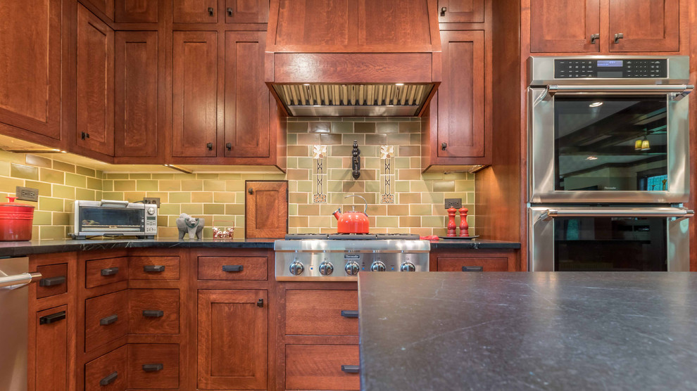 Mid-sized arts and crafts l-shaped open plan kitchen in Chicago with an undermount sink, shaker cabinets, medium wood cabinets, soapstone benchtops, green splashback, ceramic splashback, stainless steel appliances, light hardwood floors, with island, black benchtop, brown floor and wallpaper.