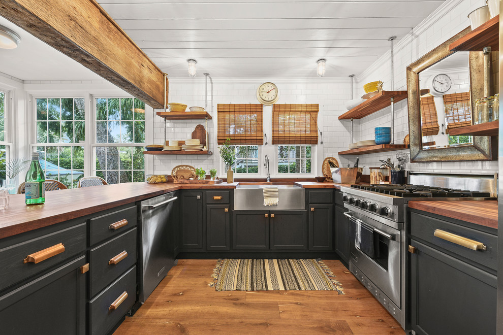 Beach style u-shaped kitchen in Jacksonville with a farmhouse sink, recessed-panel cabinets, black cabinets, wood benchtops, white splashback, subway tile splashback, stainless steel appliances, medium hardwood floors, a peninsula, brown floor and brown benchtop.