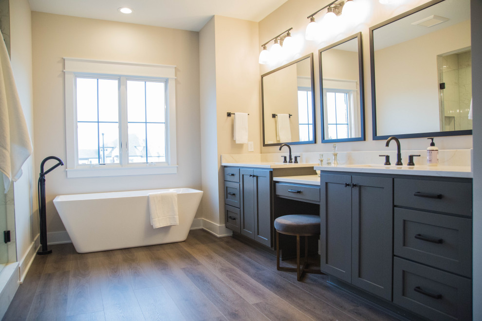 Large traditional ensuite bathroom in Indianapolis with recessed-panel cabinets, grey cabinets, a freestanding bath, an alcove shower, a two-piece toilet, beige walls, medium hardwood flooring, a submerged sink, granite worktops, brown floors, a hinged door, white worktops, double sinks and a built in vanity unit.