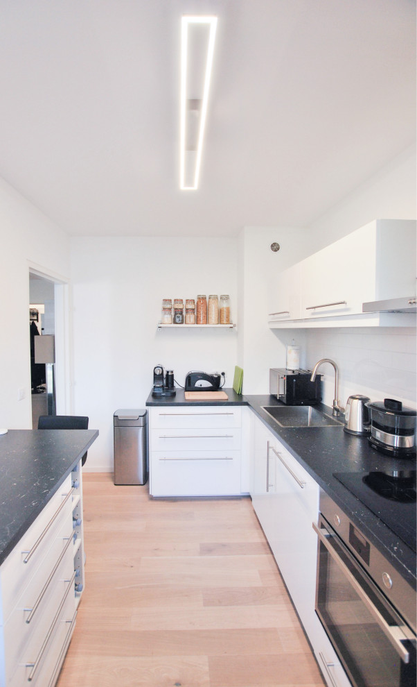This is an example of a mid-sized contemporary l-shaped separate kitchen in Other with a single-bowl sink, flat-panel cabinets, white cabinets, laminate benchtops, white splashback, subway tile splashback, stainless steel appliances, light hardwood floors, with island and black benchtop.