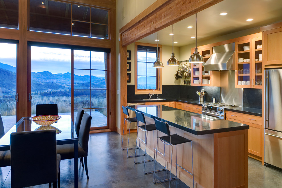 Inspiration for a mid-sized country l-shaped eat-in kitchen in Seattle with stainless steel appliances, glass-front cabinets, medium wood cabinets, black splashback, stone slab splashback, an undermount sink, quartzite benchtops, concrete floors, with island and black benchtop.