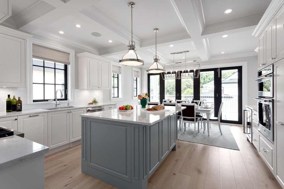 Design ideas for a large traditional eat-in kitchen in Vancouver with light hardwood floors, brown floor, a double-bowl sink, recessed-panel cabinets, blue cabinets, quartz benchtops, white splashback, porcelain splashback, stainless steel appliances, with island and white benchtop.
