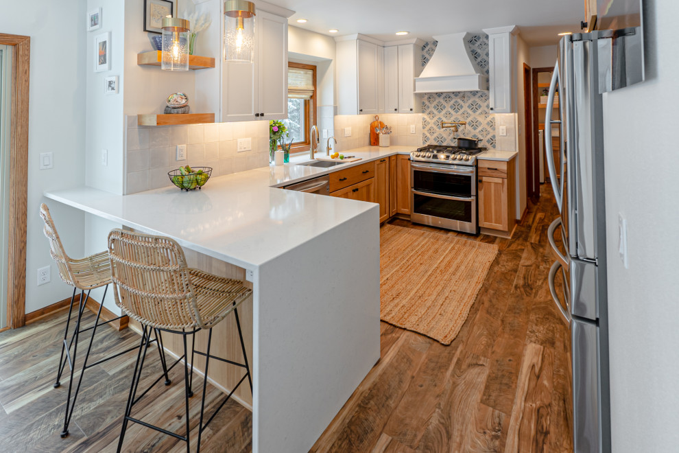 Photo of a mid-sized beach style u-shaped eat-in kitchen in Milwaukee with an undermount sink, shaker cabinets, white cabinets, quartz benchtops, beige splashback, ceramic splashback, stainless steel appliances, medium hardwood floors, a peninsula, brown floor and white benchtop.