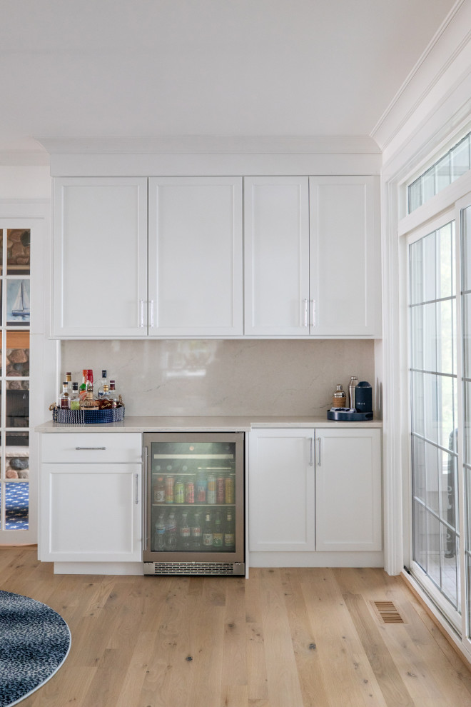 This is an example of a small beach style single-wall home bar in Milwaukee with recessed-panel cabinets, white cabinets, beige splashback, engineered quartz splashback, light hardwood floors and beige benchtop.