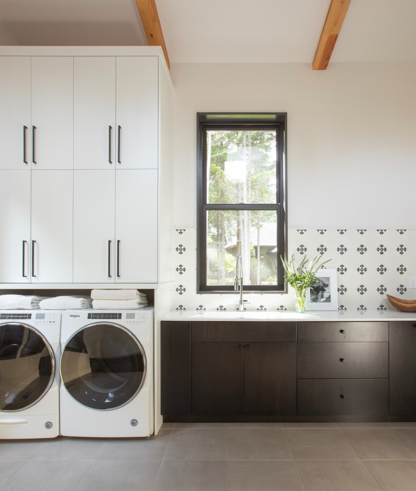 Photo of a mid-sized modern single-wall dedicated laundry room in Seattle with an undermount sink, flat-panel cabinets, black cabinets, solid surface benchtops, black splashback, cement tile splashback, white walls, porcelain floors, a side-by-side washer and dryer, grey floor, white benchtop and exposed beam.