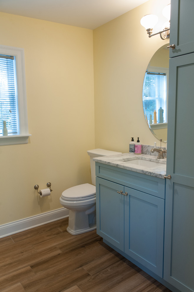 This is an example of an eclectic powder room in Philadelphia with recessed-panel cabinets, blue cabinets, a two-piece toilet, yellow walls, porcelain floors, an undermount sink, marble benchtops and brown floor.