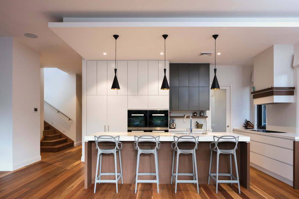 Photo of a contemporary l-shaped kitchen in Sydney with an undermount sink, flat-panel cabinets, white cabinets, black appliances, medium hardwood floors, with island, brown floor and white benchtop.