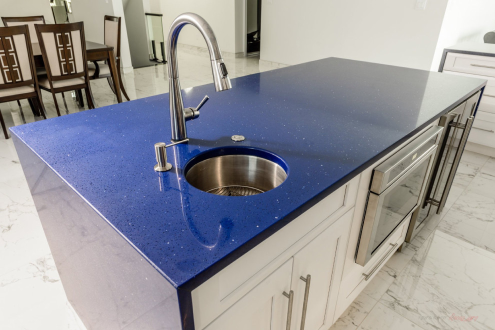 Large modern l-shaped eat-in kitchen in New York with a farmhouse sink, recessed-panel cabinets, white cabinets, quartz benchtops, black splashback, engineered quartz splashback, stainless steel appliances, marble floors, with island, multi-coloured floor and blue benchtop.