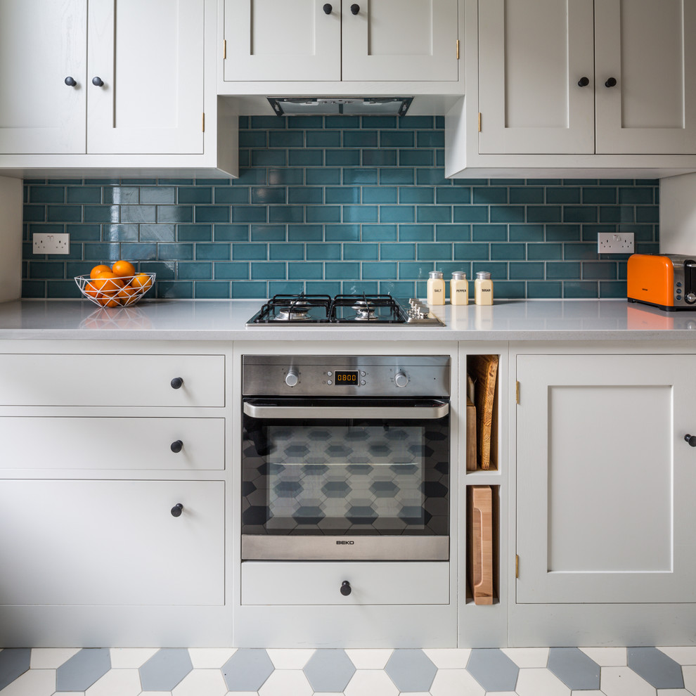 This is an example of a small contemporary l-shaped open plan kitchen in London with a farmhouse sink, shaker cabinets, white cabinets, quartz benchtops, blue splashback, subway tile splashback, stainless steel appliances, ceramic floors, a peninsula and multi-coloured floor.