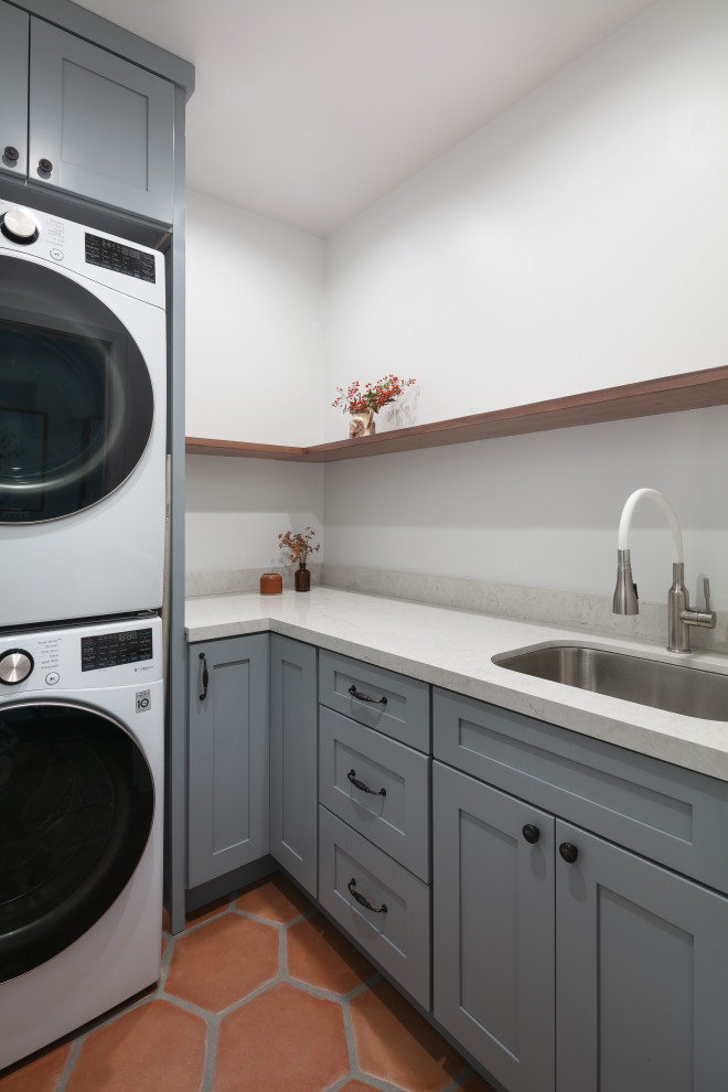This is an example of a small classic l-shaped separated utility room in Los Angeles with a submerged sink, shaker cabinets, grey cabinets, engineered stone countertops, white splashback, stone slab splashback, white walls, terracotta flooring, a stacked washer and dryer, orange floors and white worktops.