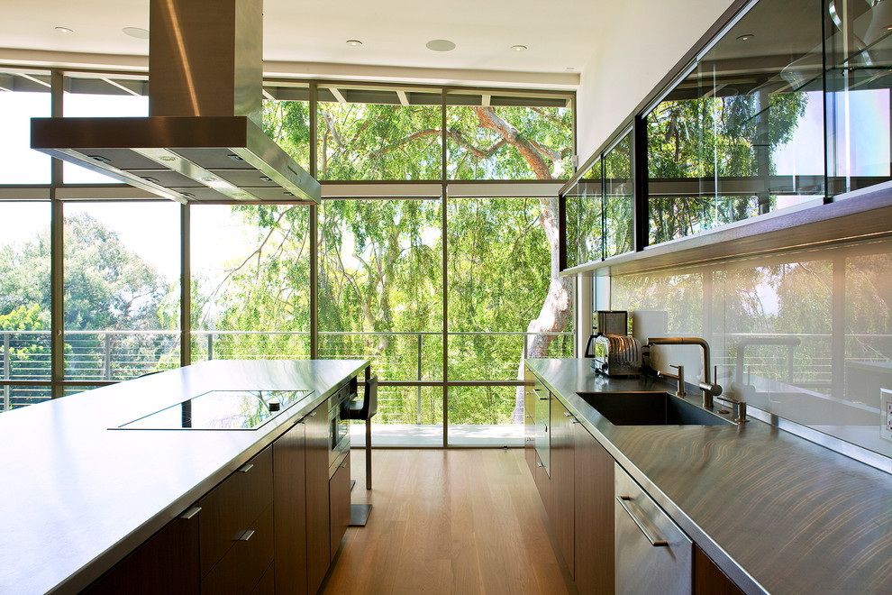 Photo of a midcentury galley kitchen in Los Angeles with an undermount sink, flat-panel cabinets, dark wood cabinets, stainless steel benchtops, white splashback, glass sheet splashback and medium hardwood floors.