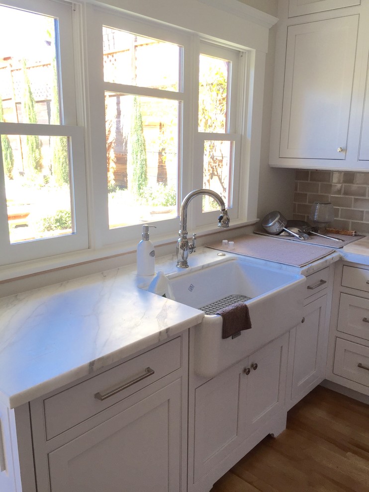 Small arts and crafts u-shaped eat-in kitchen in San Diego with a farmhouse sink, shaker cabinets, white cabinets, marble benchtops, beige splashback, subway tile splashback, stainless steel appliances, light hardwood floors and a peninsula.