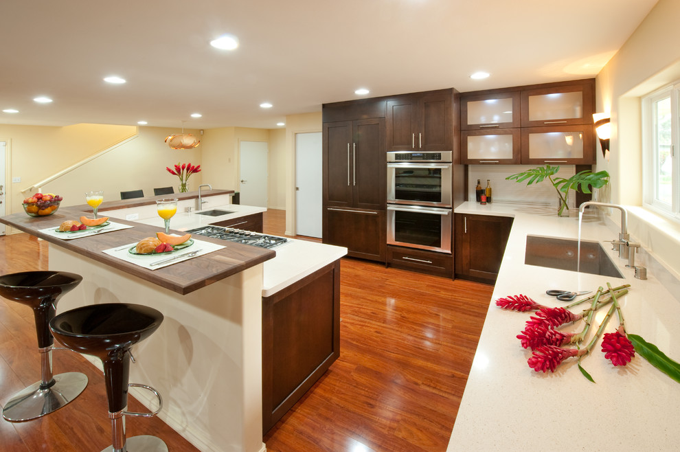 Large tropical l-shaped kitchen in Hawaii with an undermount sink, shaker cabinets, dark wood cabinets, solid surface benchtops, white splashback, panelled appliances, medium hardwood floors, no island and brown floor.