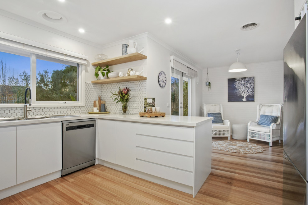 Photo of a mid-sized beach style u-shaped eat-in kitchen in Geelong with a double-bowl sink, white cabinets, soapstone benchtops, white splashback, ceramic splashback, stainless steel appliances, light hardwood floors and with island.