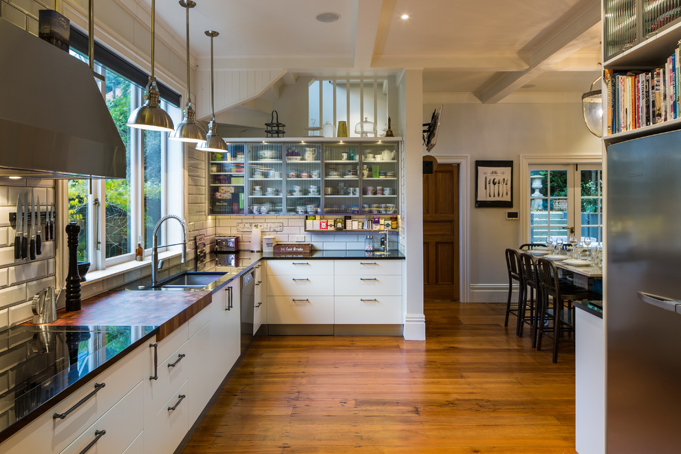 Photo of a large industrial l-shaped eat-in kitchen in Wellington with an undermount sink, glass-front cabinets, white cabinets, granite benchtops, white splashback, ceramic splashback, stainless steel appliances, medium hardwood floors and with island.