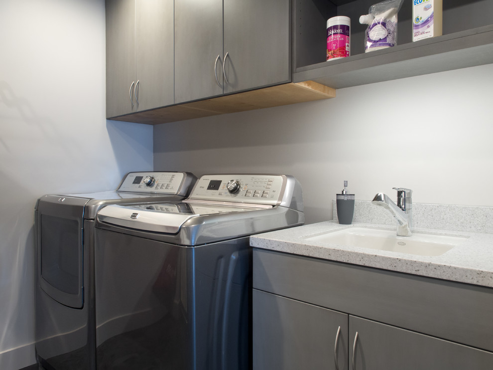 Design ideas for a mid-sized contemporary galley dedicated laundry room in Seattle with flat-panel cabinets, grey cabinets, solid surface benchtops, grey walls, concrete floors, a side-by-side washer and dryer, an undermount sink, grey splashback, black floor and grey benchtop.